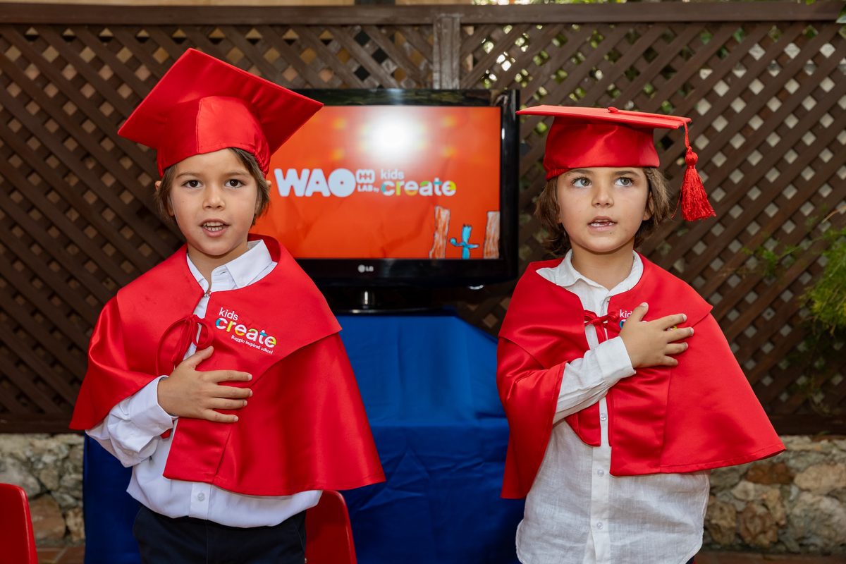 Dos niños graduados con toga y birrete rojos frente a la pantalla de WAO Lab by Kids Create